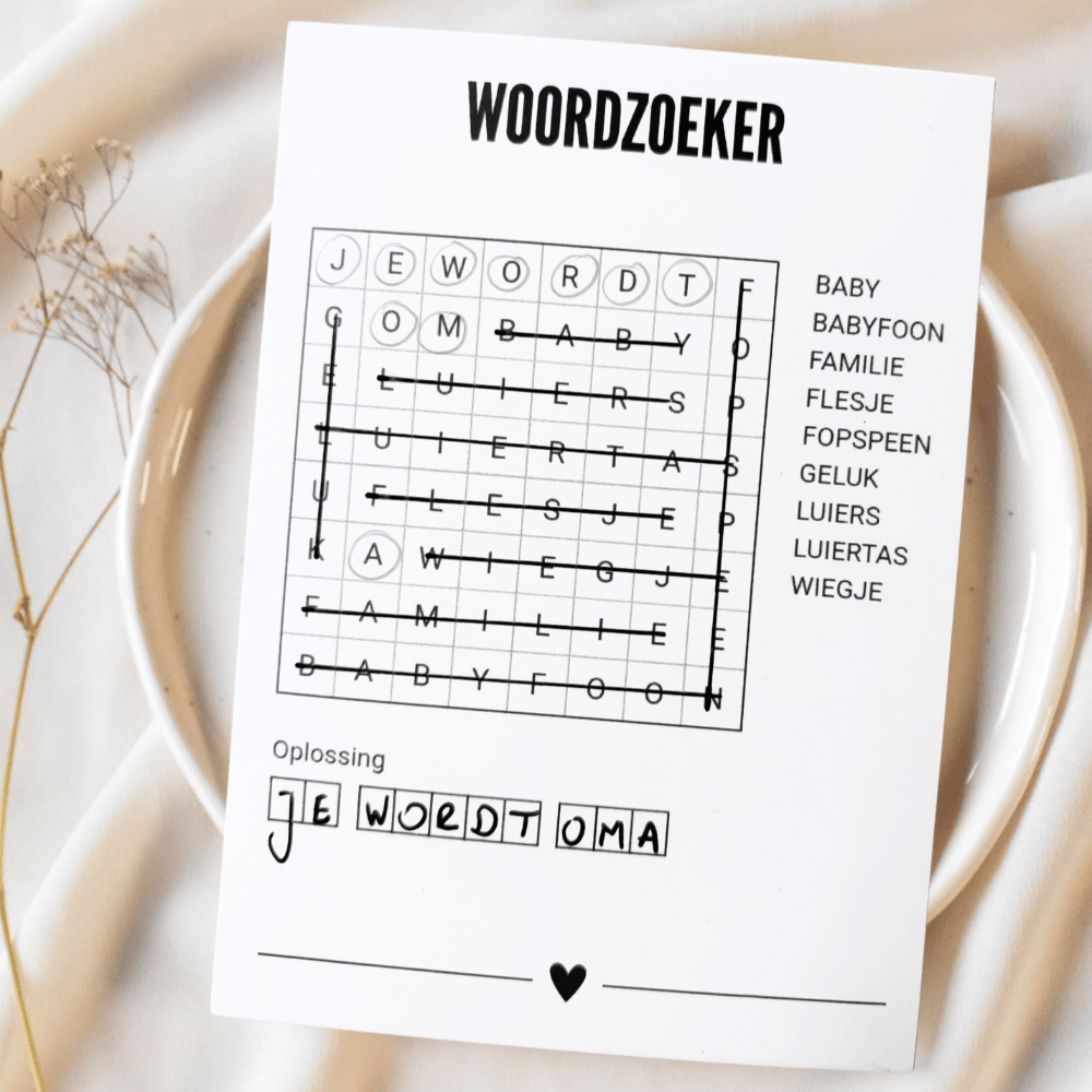A6 woordzoekerkaart met doorgestreepte woorden en de oplossing 'Je wordt oma', op een bord met lichte stof eromheen.