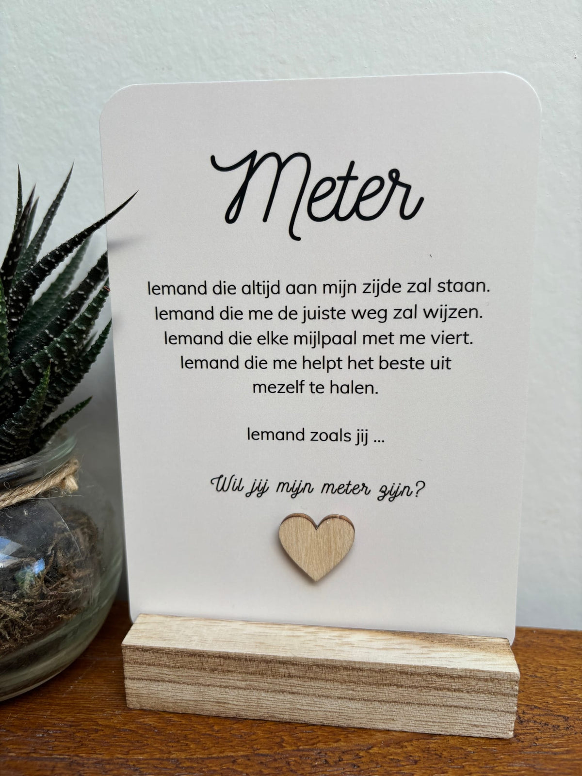 Witte ansichtkaart met tekst ‘Wil jij mijn meter zijn?’ en houten hartje, in een houten houder naast een plant.