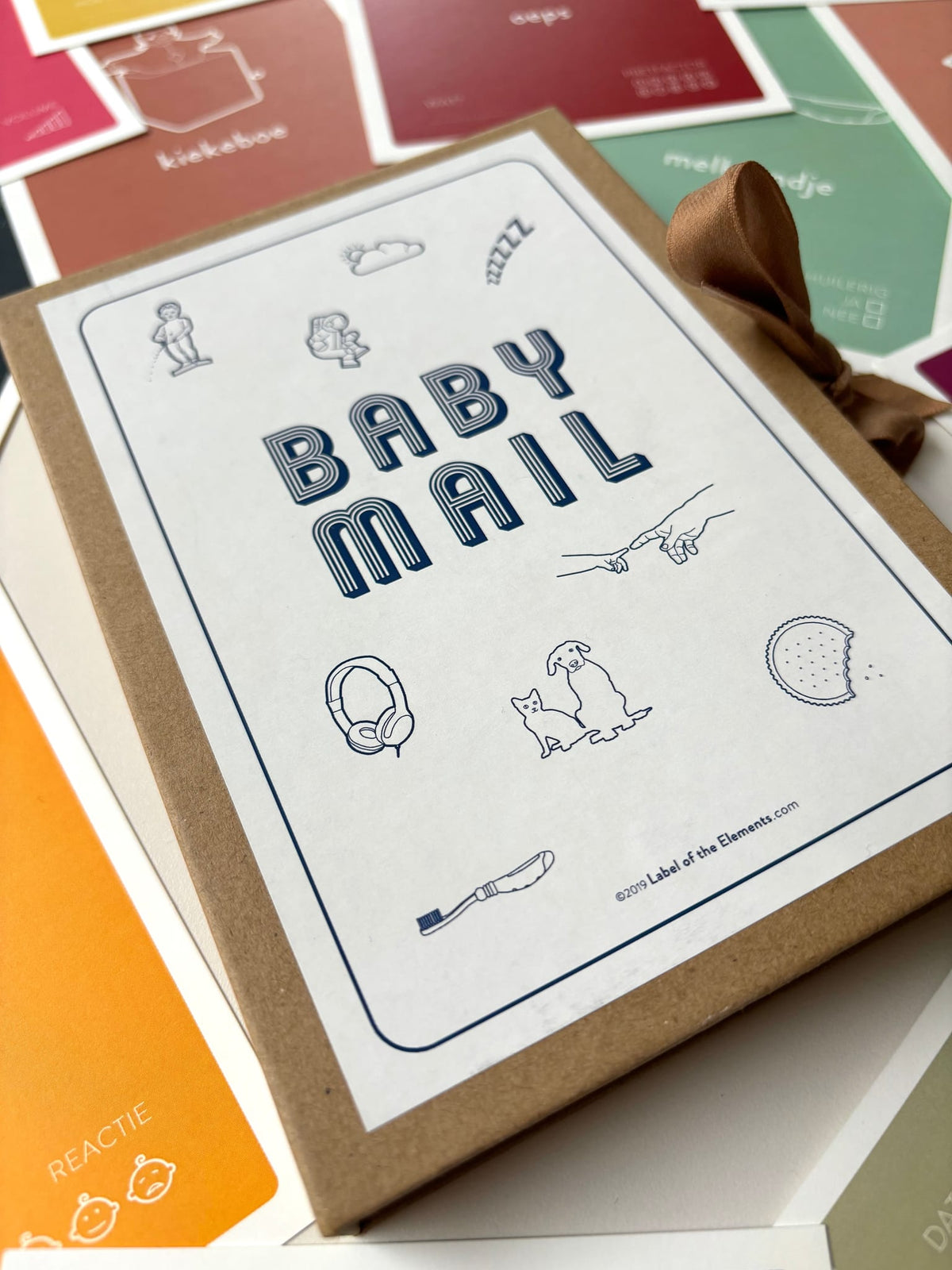 Baby Mail-mijlpaalkaarten in een bruin bewaardoosje met sluitlint, omringd door gekleurde kaarten.