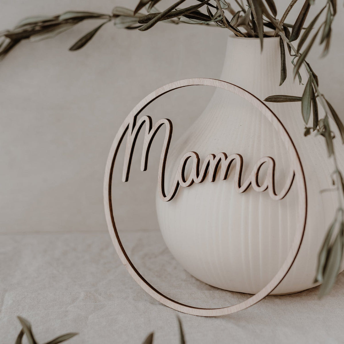 Lichtbruine houten krans met het woord mama leunt tegen een witte vaas met groene takken.