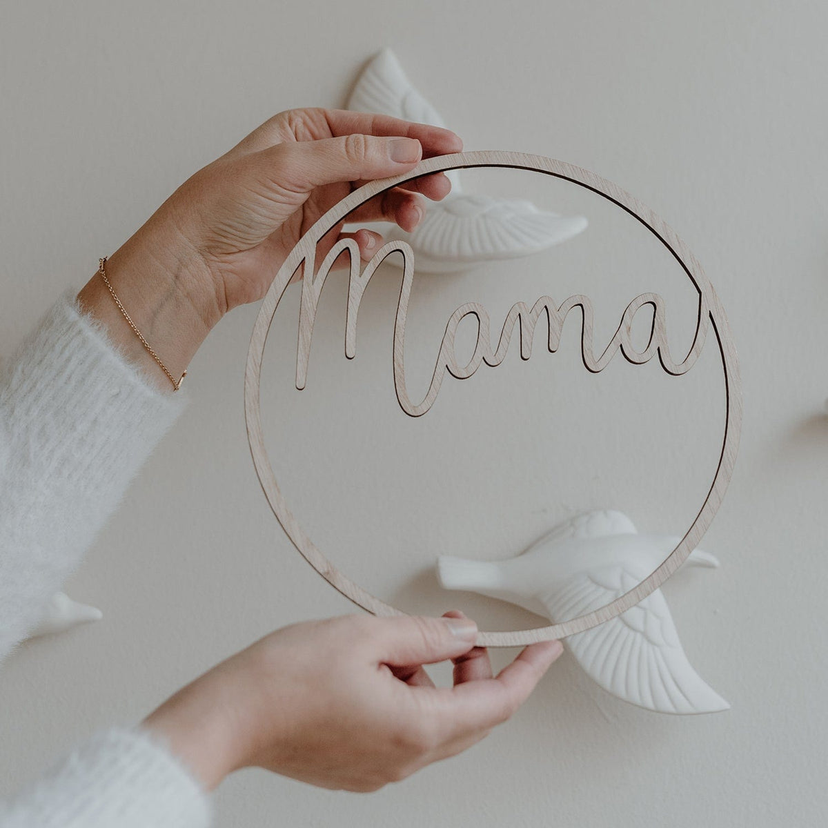 Twee handen houden een lichtbruine houten krans met het woord mama vast, met witte vogeldecoraties op de achtergrond.