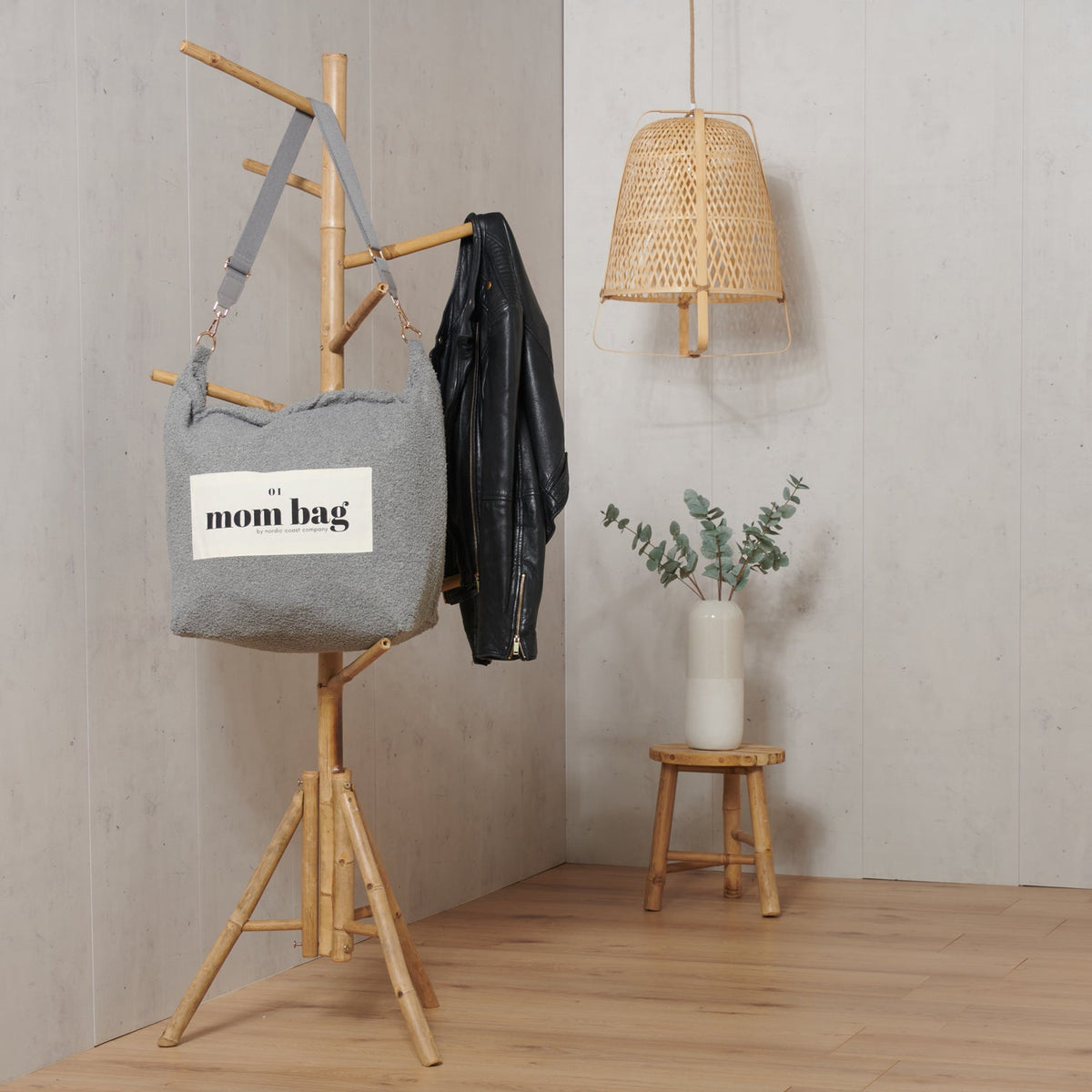 Grijze Mom Bag Teddy Bouclé met etiket 'mom bag', hangend aan een houten kapstok naast een zwarte jas.