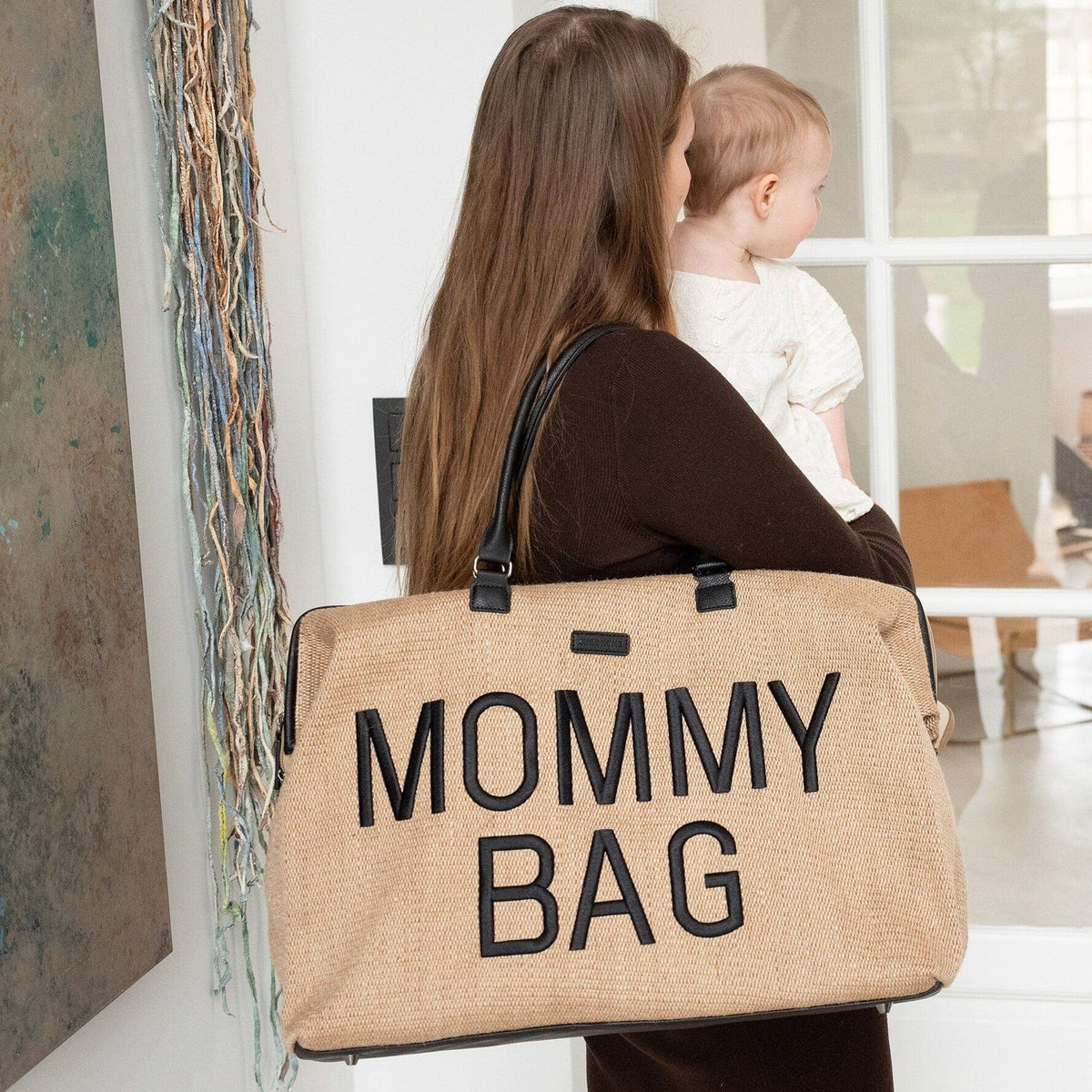 Vrouw met een kind op de arm en een beige Mommy Bag verzorgingstas met zwarte letters over haar schouder.