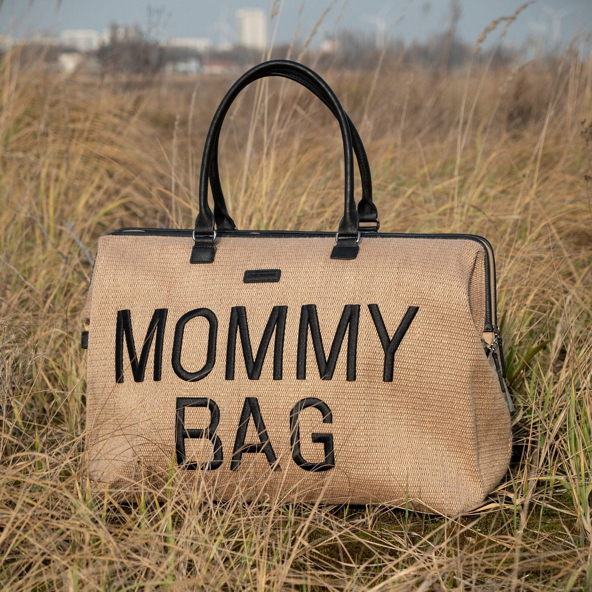 Beige Mommy Bag verzorgingstas met raffialook, zwarte handvatten en zwarte letters, buiten in het hoge gras.