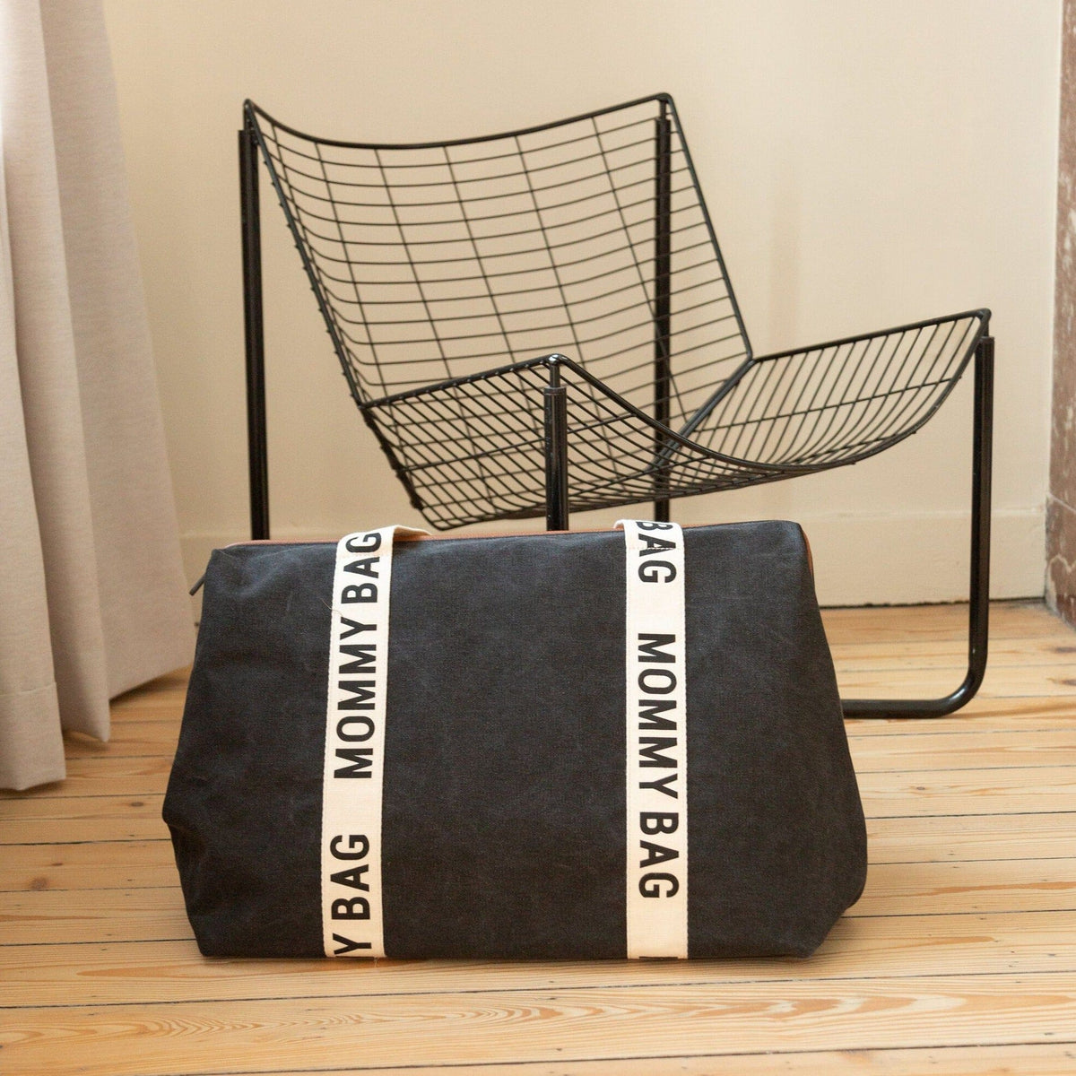 Zwarte Mommy Bag verzorgingstas met crèmekleurige banden op een houten vloer voor een zwarte stoel.