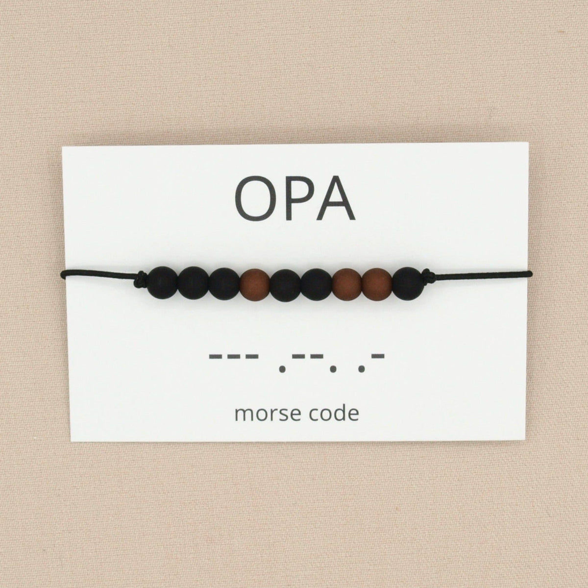 Opa-armband in morsecode met zwarte en bruine kralen aan een zwart koord, op een wit kaartje met ‘opa’ en morsecode.