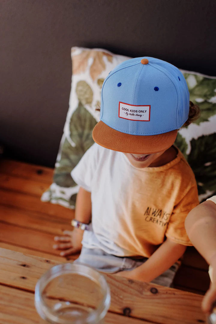 Kind aan een houten tafel met de blauwe Mini Jean pet van Hello Hossy, met de bruine klep naar voren.