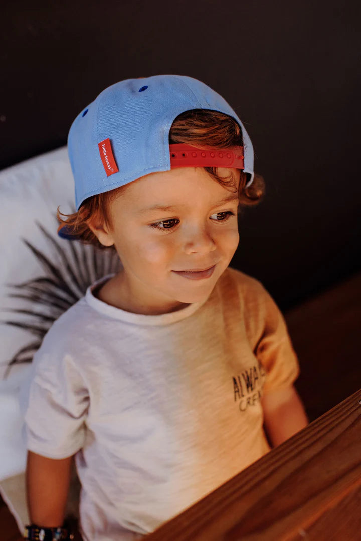 Kind draagt de blauwe Mini Jean pet van Hello Hossy achterstevoren, met de rode verstelbare sluiting in beeld.