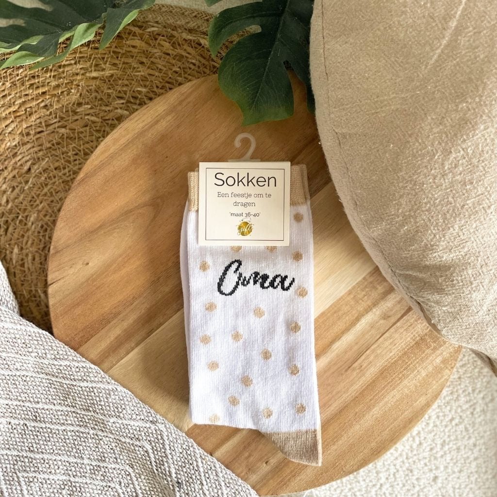 Witte sokken met beige stippen en het opschrift ‘Oma’, met productlabel op een ronde houten plank.