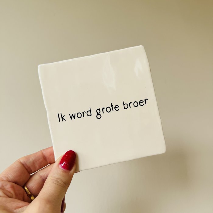 Wit tegeltje met de zwarte tekst 'Ik word grote broer', vastgehouden in een hand tegen een beige achtergrond.