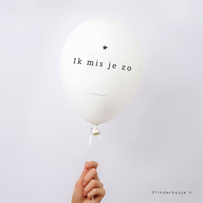 Witte troostballon met de tekst 'Ik mis je zo' en een zwart sterretje, aan een lint vastgehouden door een hand.