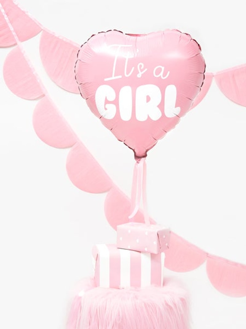 Roze hartvormige folieballon 'It's a Girl' boven roze cadeaudoosjes, met roze slingers op de achtergrond.