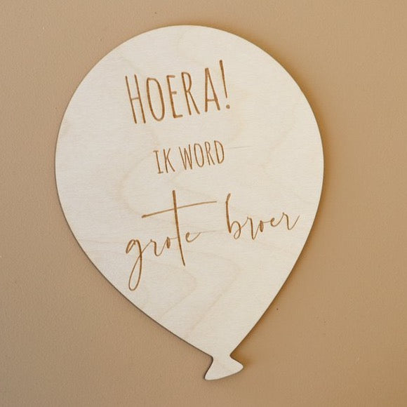 Lichtgekleurde houten ballon met de tekst 'Hoera! Ik word grote broer' op een bruine achtergrond.