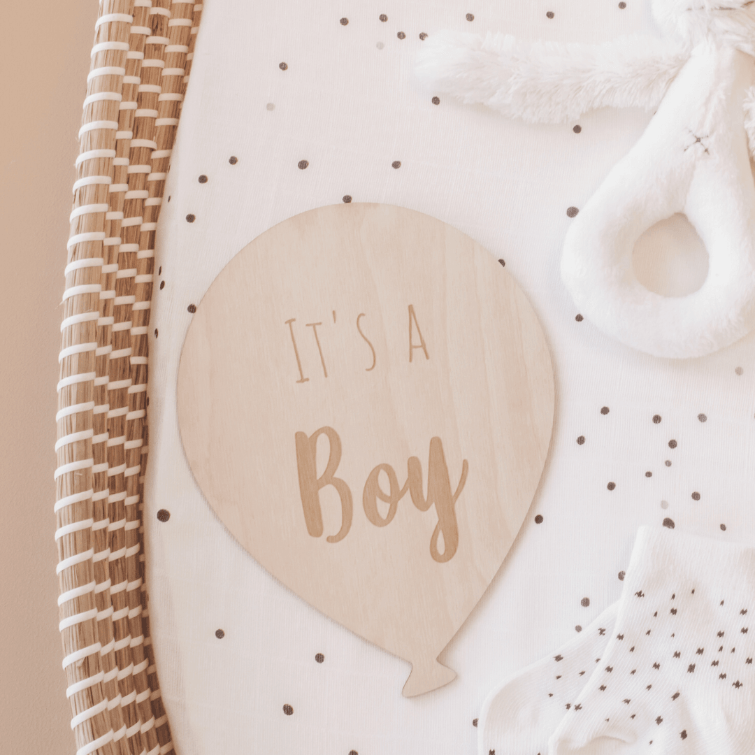 Houten ballon met opdruk ‘It's a boy’ op een gestippeld matras in een rieten mand, naast een witte knuffel.