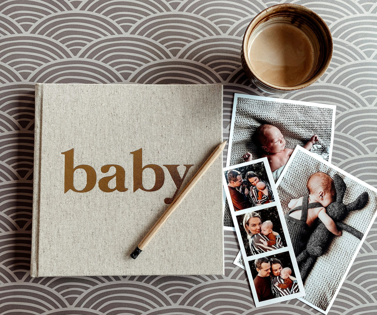 Linnen baby-invulboek met naturelkleurige kaft en gouden tekst 'baby', naast babyfoto's en een potlood.