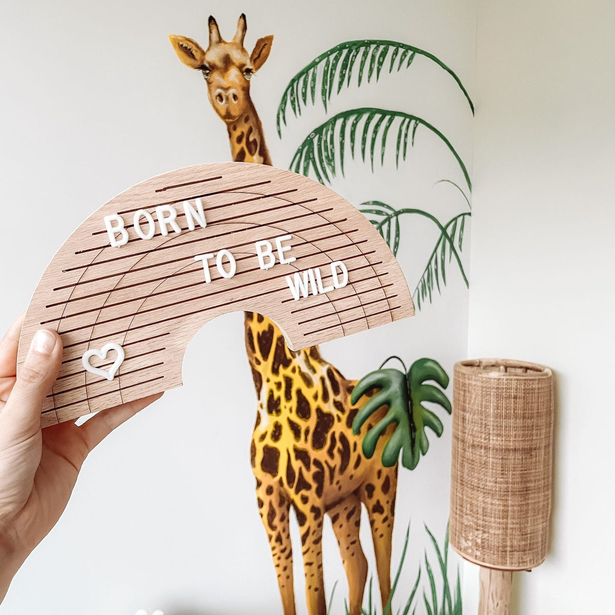 Houten regenboogletterbord met 'born to be wild' en een hartje, vastgehouden voor een giraf op de muur.
