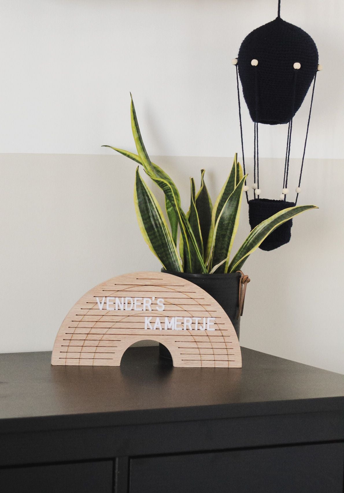 Houten regenboogletterbord met witte letters op een donkere kast, naast een plant en een zwarte luchtballondecoratie.