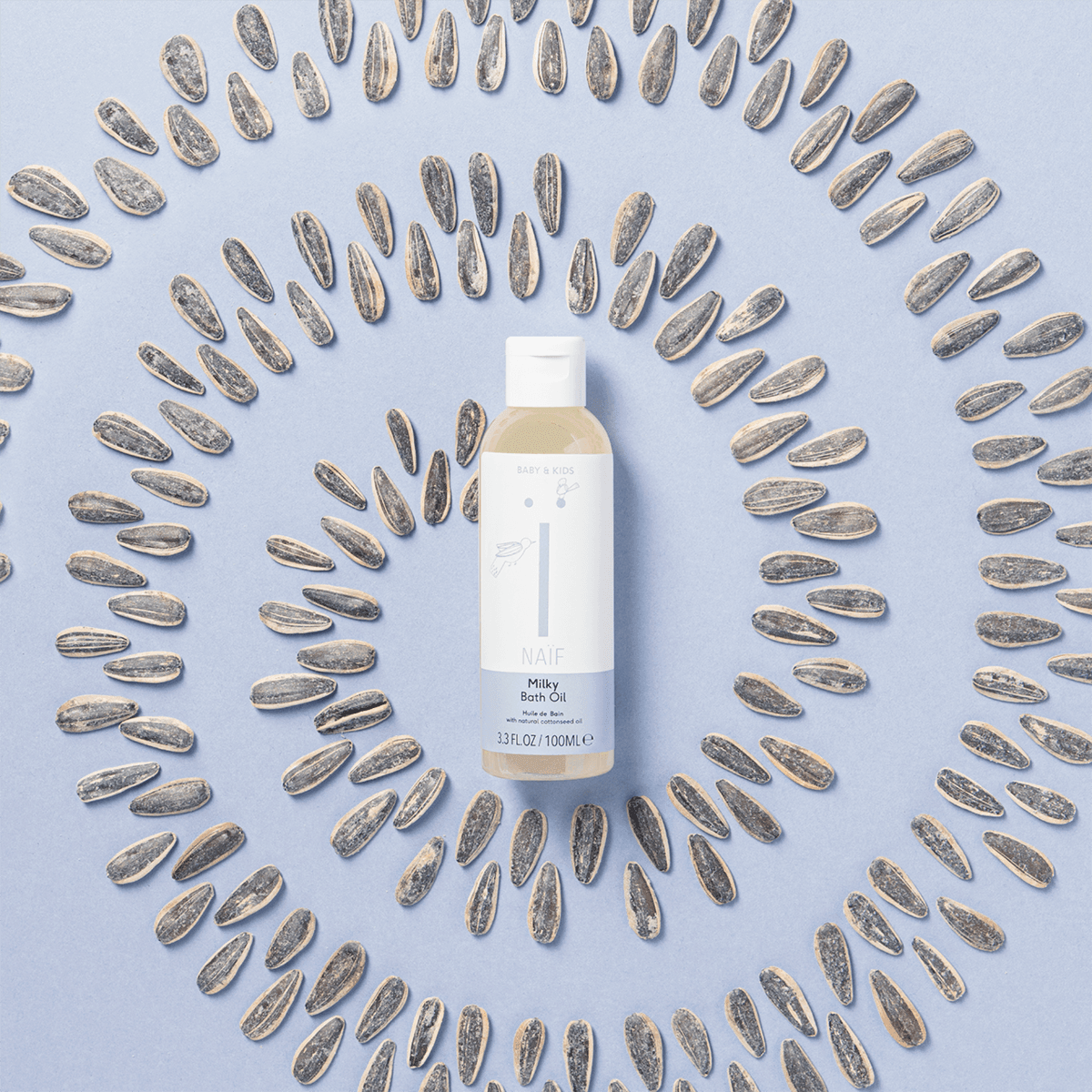 Fles Naïf Milky Badolie omringd door zonnebloempitten in cirkels op een lichtblauwe achtergrond