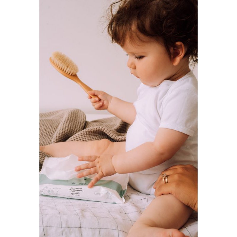 Baby met een haarborstel in de hand zit naast een geopend pak Naïf plasticvrije billendoekjes.