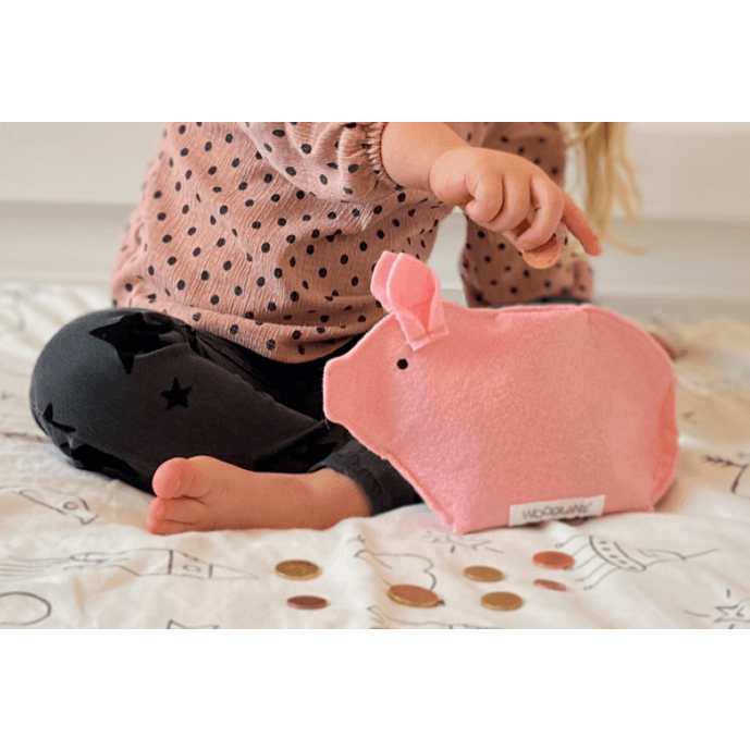 Kind houdt een munt boven het roze Piggy-spaarvarken, met losse munten op het bed