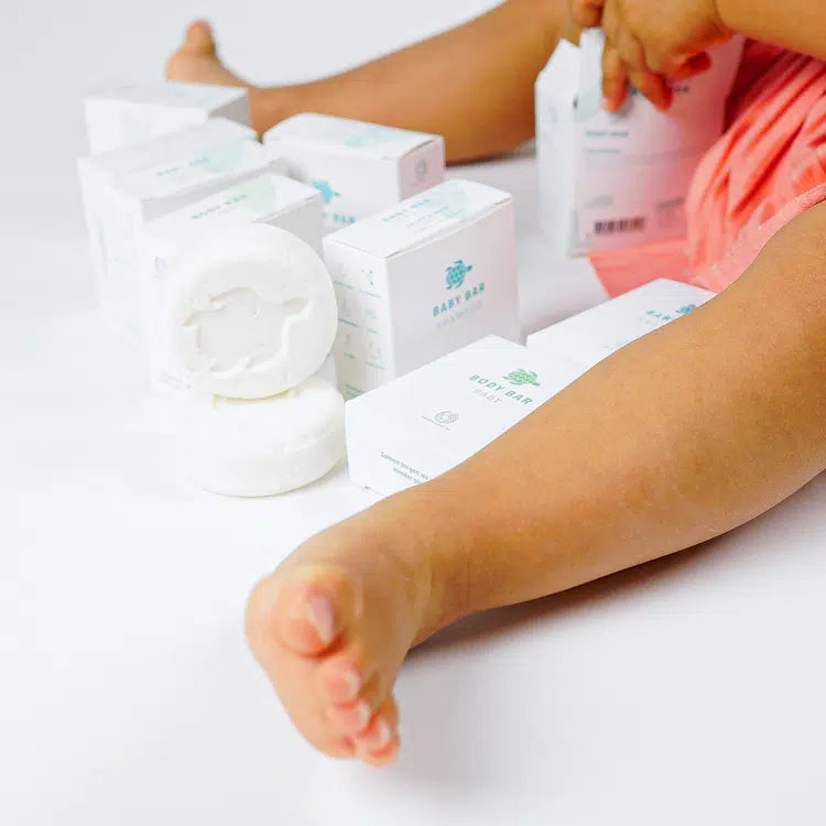 Zittend kind met Shampoo Bar Baby-verpakkingen en twee witte shampoobars, waarvan één met zichtbaar schildpadmotief.