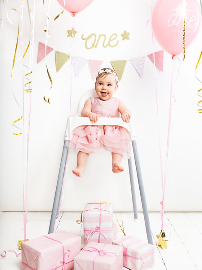 Roze slinger boven een baby in een kinderstoel, tussen roze ballonnen en cadeautjes.