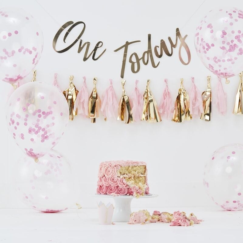 Roze cake smash kit met confettiballonnen, kwastjesslinger en gouden 'One Today'-slinger achter een aangesneden taart.