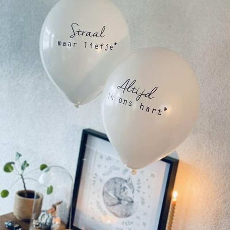 Witte troostballonnen 'Straal maar liefje' en 'Altijd in ons hart' boven een ingelijste prent naast een kaars.