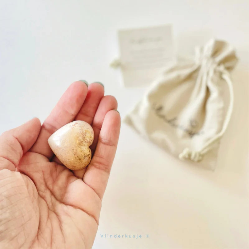 Hand met een lichtgekleurd spekstenen knuffelhartje, met het beige zakje en minikaartje op de achtergrond.