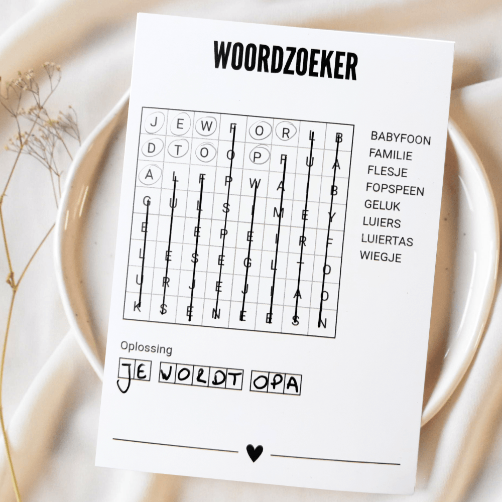 Witte A6-woordzoekerkaart met ingevulde oplossing ‘Je wordt opa’ op een bord met lichte stof en droogbloemen.