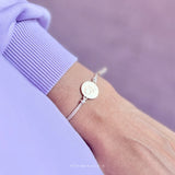 Armbandje met lichtroze koord en ronde goudkleurige vlinderbedel om een pols bij een lila mouw.