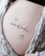 Zwangere buik met de plaktattoo ‘Little Wonder’ en twee hartjes, omlijst door een grijs gebreid vest.
