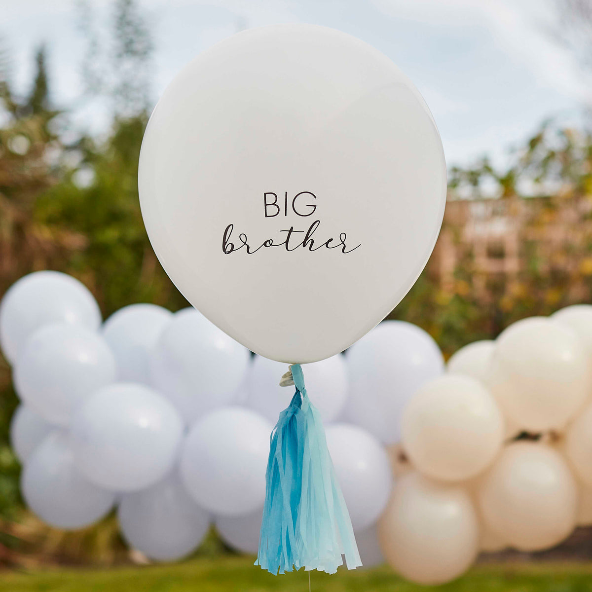 Crèmekleurige ballon met tekst 'Big brother' en blauwe kwastjes, buiten voor een slinger van ballonnen.
