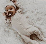 Baby in beige boxpakje met ribstructuur en bijpassend mutsje, liggend op een wit, pluizig kleed.