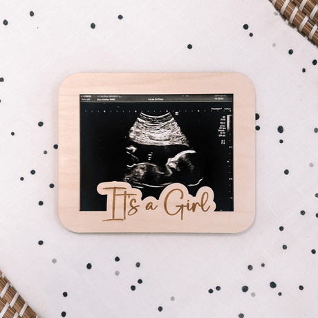 Licht houten echolijstje met echofoto en de tekst ‘It's a Girl’ op een witte ondergrond met donkere stippen.