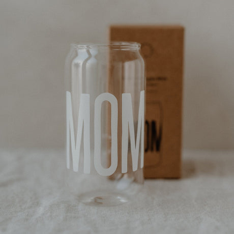 Hoog transparant drinkglas met wit opschrift ‘Mom’, met een bruine doos op de achtergrond.