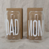 Twee hoge transparante drinkglazen met witte opschriften ‘Dad’ en ‘Mom’, met bruine dozen erachter.
