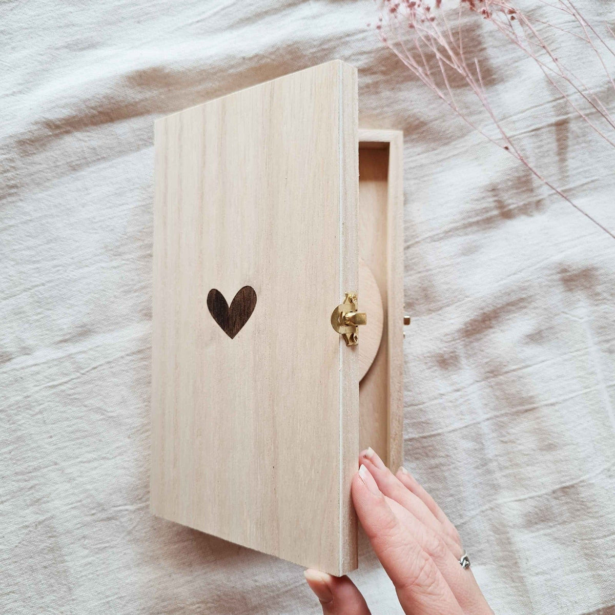 Hand opent een licht houten kistje met gegraveerd hartje en goudkleurige sluiting op een stoffen ondergrond.