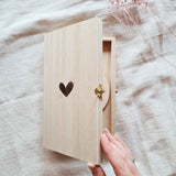 Een hand opent het houten kistje met gegraveerd hartje; de ronde houten schijf is deels zichtbaar.