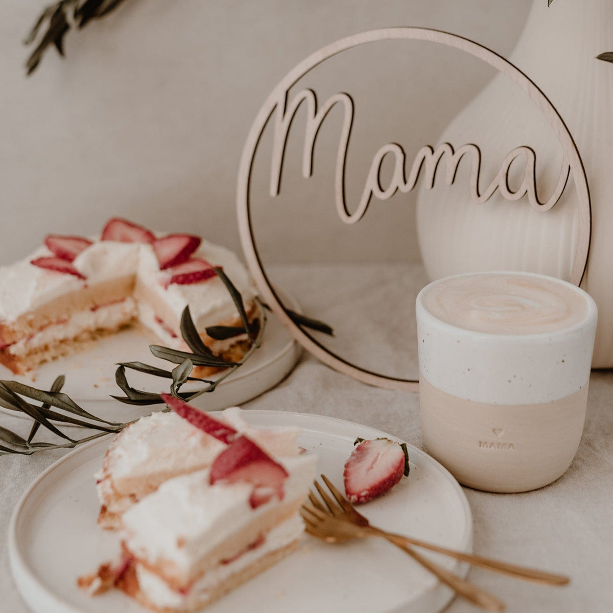 Houten krans met het woord mama naast een witte vaas, een kopje en gebak met aardbeien.