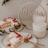 Houten krans met het woord mama naast een witte vaas, een kopje en gebak met aardbeien.