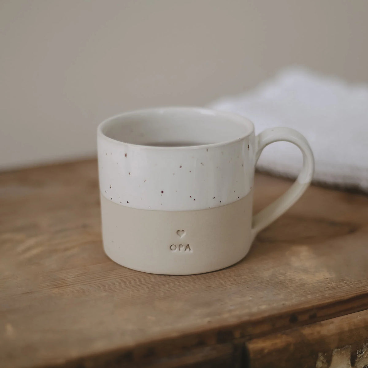 Beige koffiemok met wit gespikkelde bovenkant, oor en opschrift 'opa' op een houten tafel.