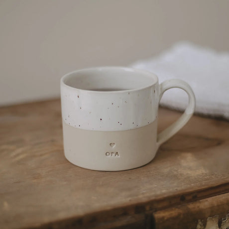 Beige koffiemok met wit gespikkelde bovenkant, oor en opschrift 'opa' op een houten tafel.