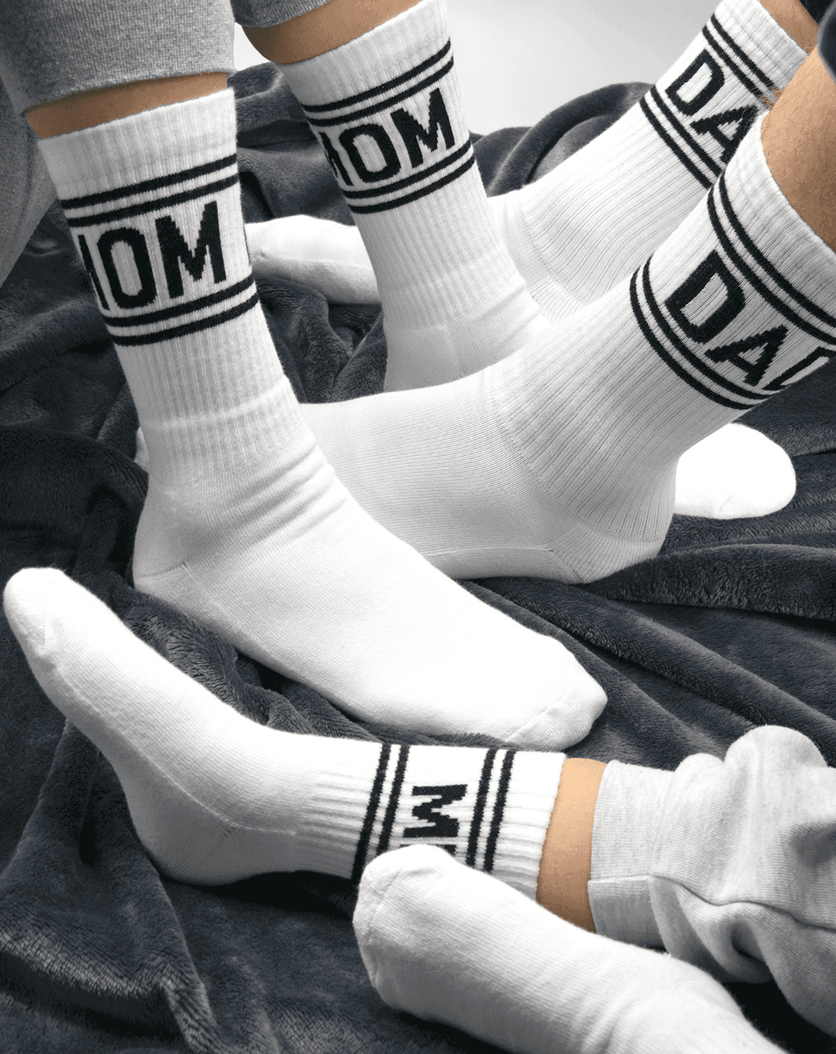 Witte tennissokken met zwarte strepen en opschriften 'mom', 'dad' en 'mini', gedragen op een donkere deken.