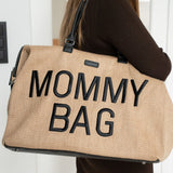 Close-up van de beige Mommy Bag verzorgingstas met zwarte letters en handvatten, gedragen over de schouder.
