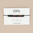 Opa-armband in morsecode met zwarte en bruine kralen aan een zwart koord, op een wit kaartje met ‘opa’ en morsecode.