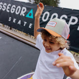 Kind met de Mini Anis pet van Hello Hossy in lichtgroen, oranje en beige, met de armen omhoog op een skatebaan.