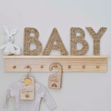 Naturelkleurige rieten letters ‘baby’ op een houten wandplank naast een wit knuffelkonijn.