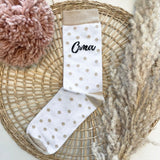 Witte sok met beige stippen en het opschrift ‘Oma’ op een gevlochten ondergrond naast droogbloemen.