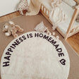 Rond crèmekleurig tapijt met zwarte tekst 'Happiness is Homemade' en hartje naast een kinderbed en knuffelbeer.